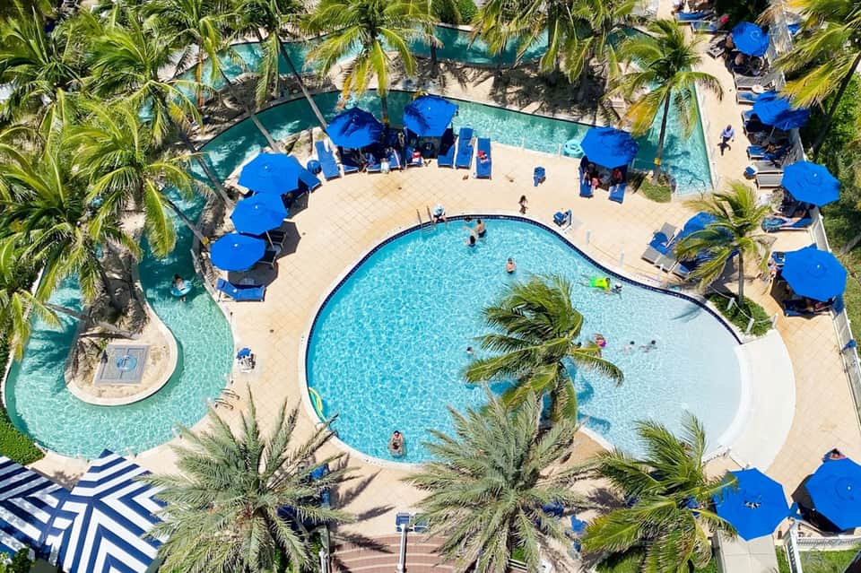 Aerial view of resort pool with blue umbrellas, palm trees, and adjacent lazy river surrounded by lounge chairs