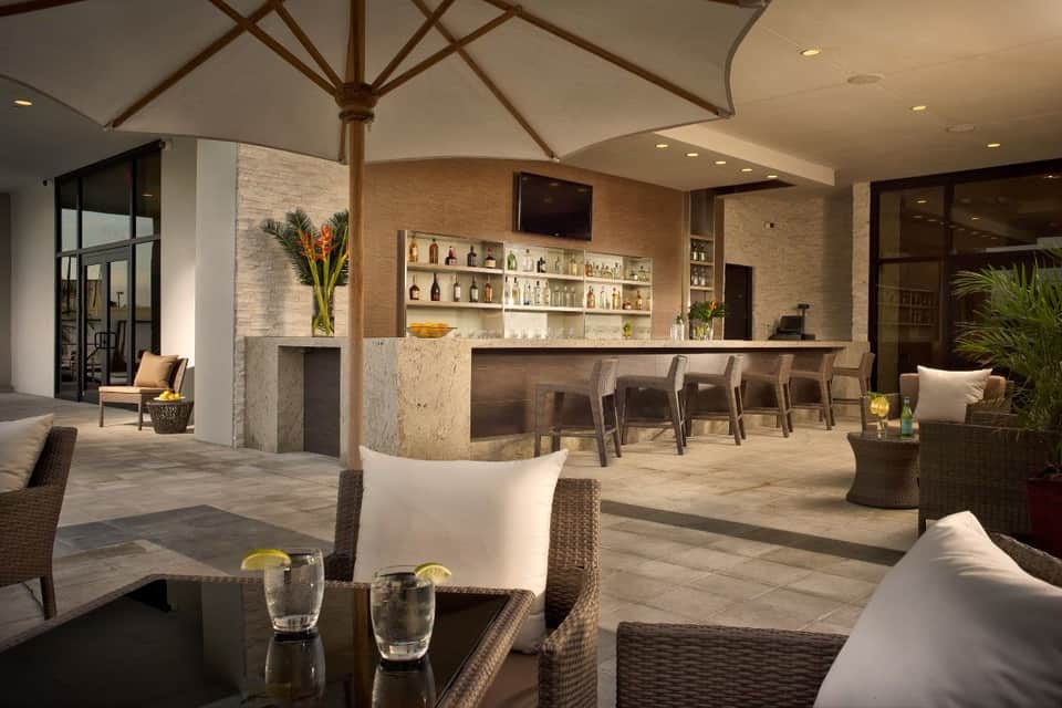 Elegant outdoor bar and lounge area with stone bar counter, wicker seating, white umbrellas, and tropical plants