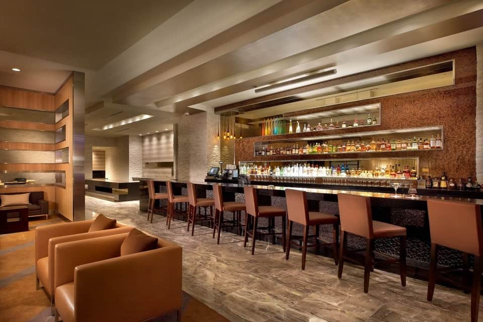Modern hotel bar interior with illuminated shelving, leather bar stools, wooden accents, and contemporary lounge seating