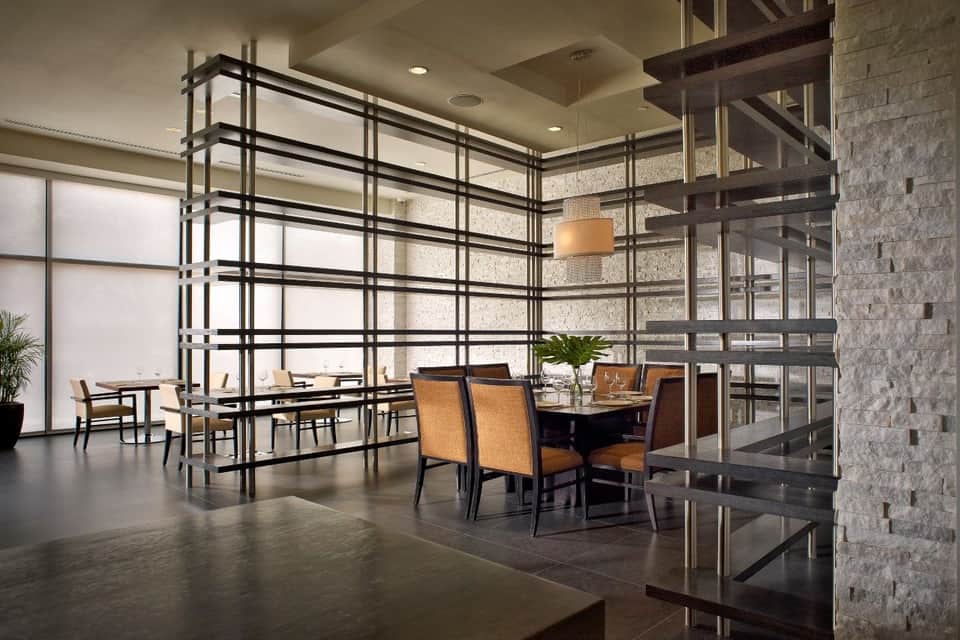 Contemporary dining space with black metal grid partition, wooden chairs, stone walls, and floor-to-ceiling windows