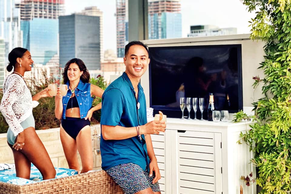 Three guests enjoying drinks on a rooftop terrace with city skyline and TV entertainment area