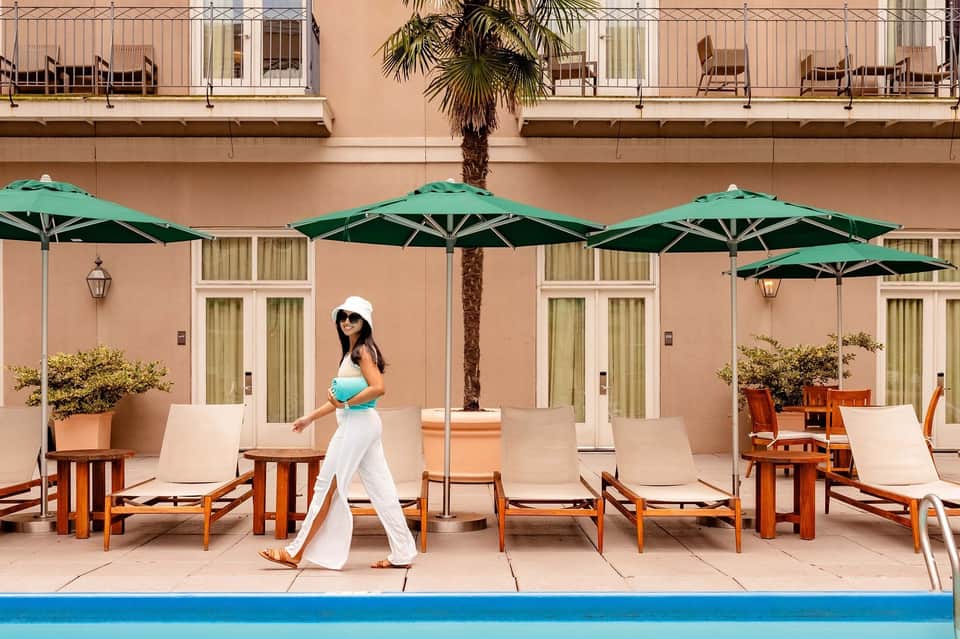 Woman in white attire walking on pool deck past green umbrellas and wooden loungers at a luxury resort with peach-colored building facade