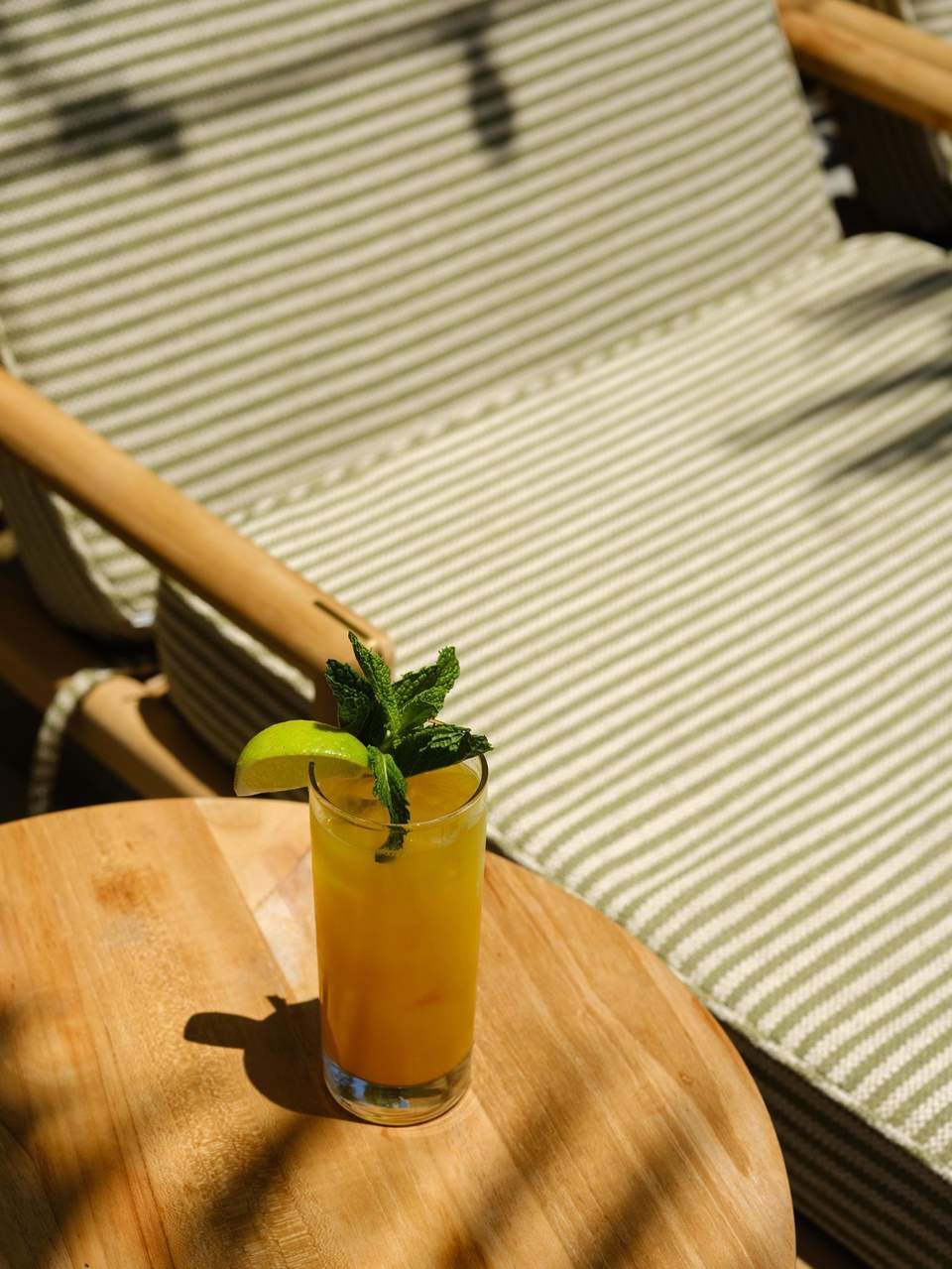 Yellow cocktail with lime and mint garnish on wooden table by striped lounge chair