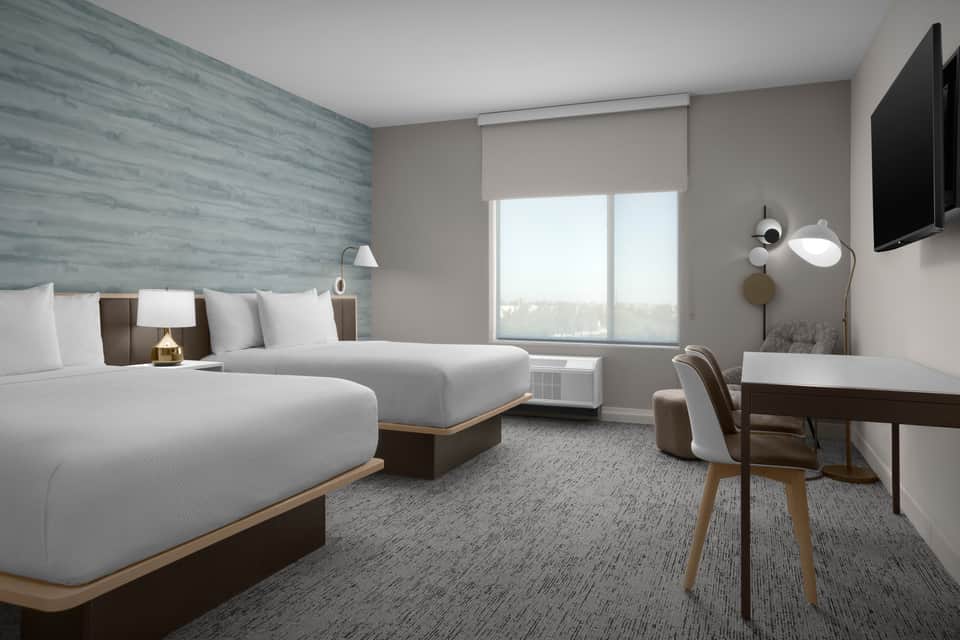 Modern hotel room with two queen beds, blue accent wall, desk, TV, and large window with city views