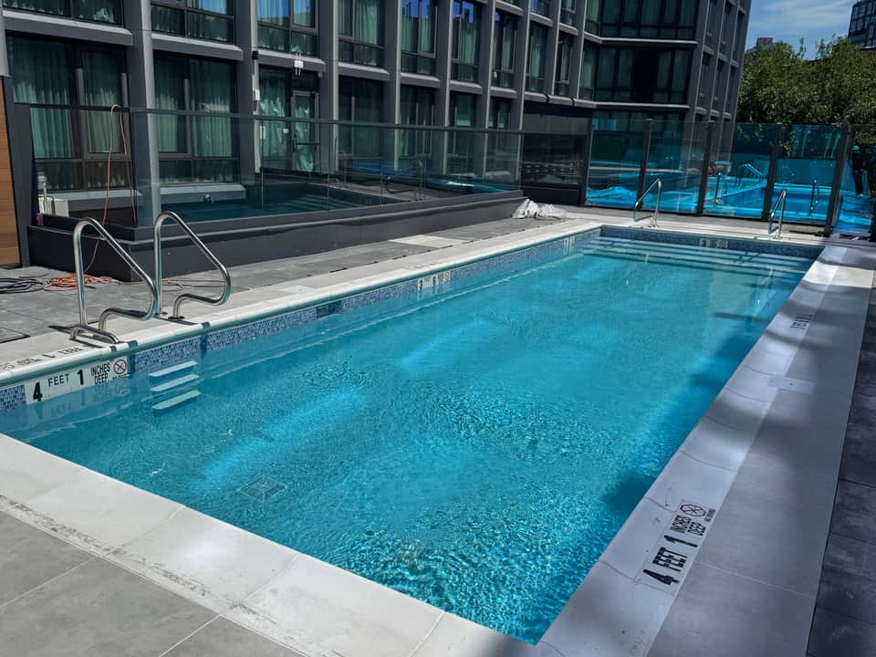 Indoor swimming pool with turquoise water, white deck, metal railings, glass barriers, and high-rise windows in background