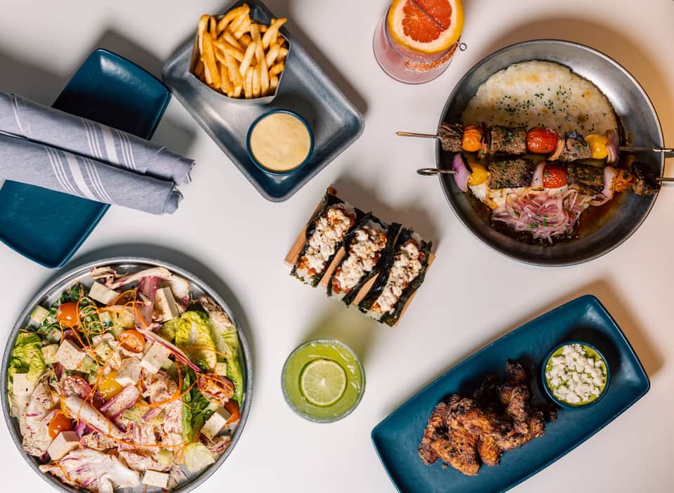 Overhead view of gourmet dishes including grilled kebabs, salad, sushi rolls, fries, and beverages on a white surface