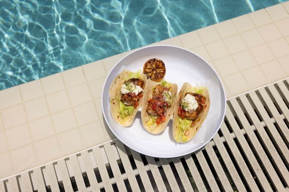 Three gourmet tacos with garnish on white plate at pool edge overlooking turquoise water