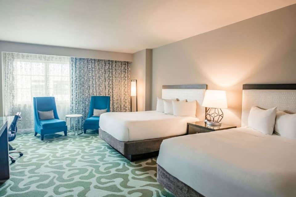 Spacious hotel room with two queen beds, blue accent chairs, patterned carpet, and modern furnishings