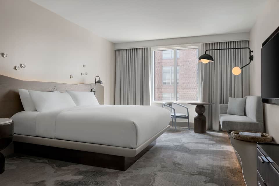 Contemporary hotel bedroom with white bedding, floor-to-ceiling windows overlooking city, neutral curtains, and modern furnishings
