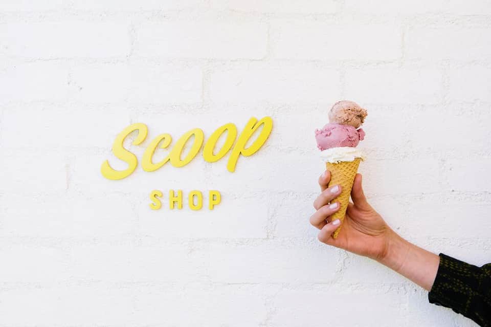 Hand holding three-scoop ice cream cone in front of yellow Scoop Shop sign on white brick wall
