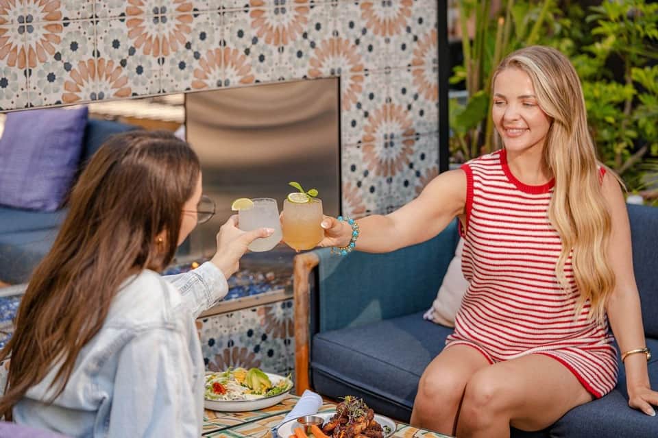 Two women toasting with cocktails at an outdoor patio with patterned tile backdrop and decorative pillows