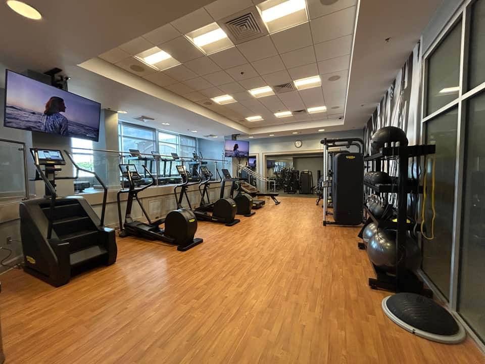 Bright fitness facility with cardio equipment, wooden floors, and large windows with nature views