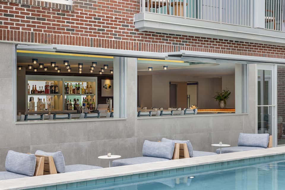 Modern poolside bar with open service window, brick backdrop, and lounge seating along pool edge