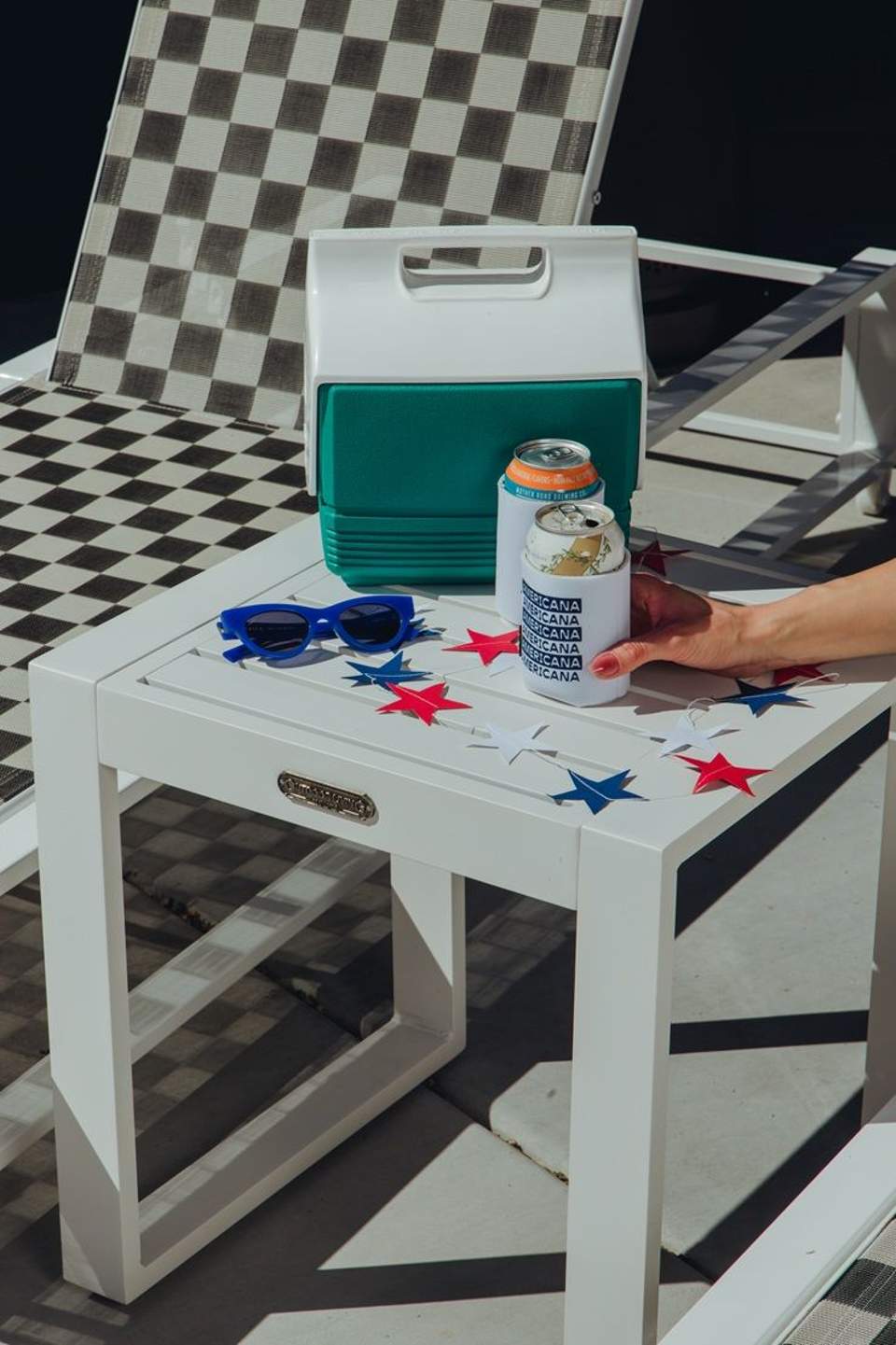 Casual outdoor seating with cooler, sunglasses, and beverage on white table with checkered pattern