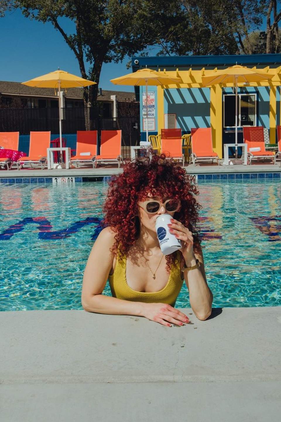Woman in yellow swimsuit at pool edge with curly red hair, white sunglasses, holding drink with yellow umbrellas and orange loungers visible