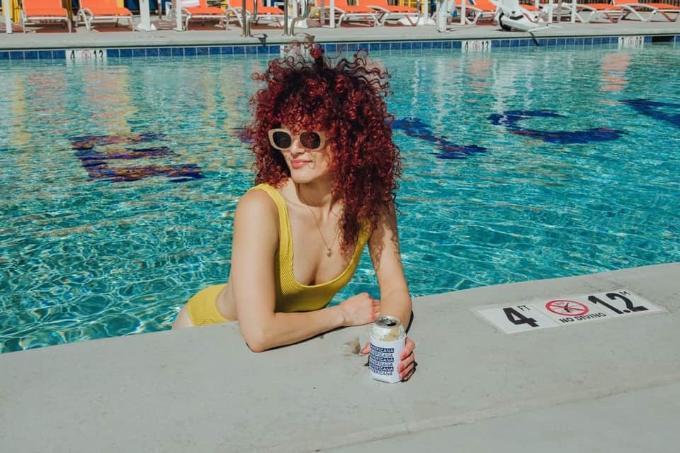 Woman in yellow swimsuit at pool deck holding beverage with lane dividers visible in clear turquoise water behind her
