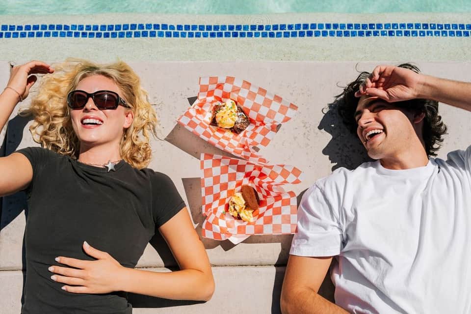 Two people in casual clothing lying on pool deck with red checkered food wrapper and items between them, pool visible behind