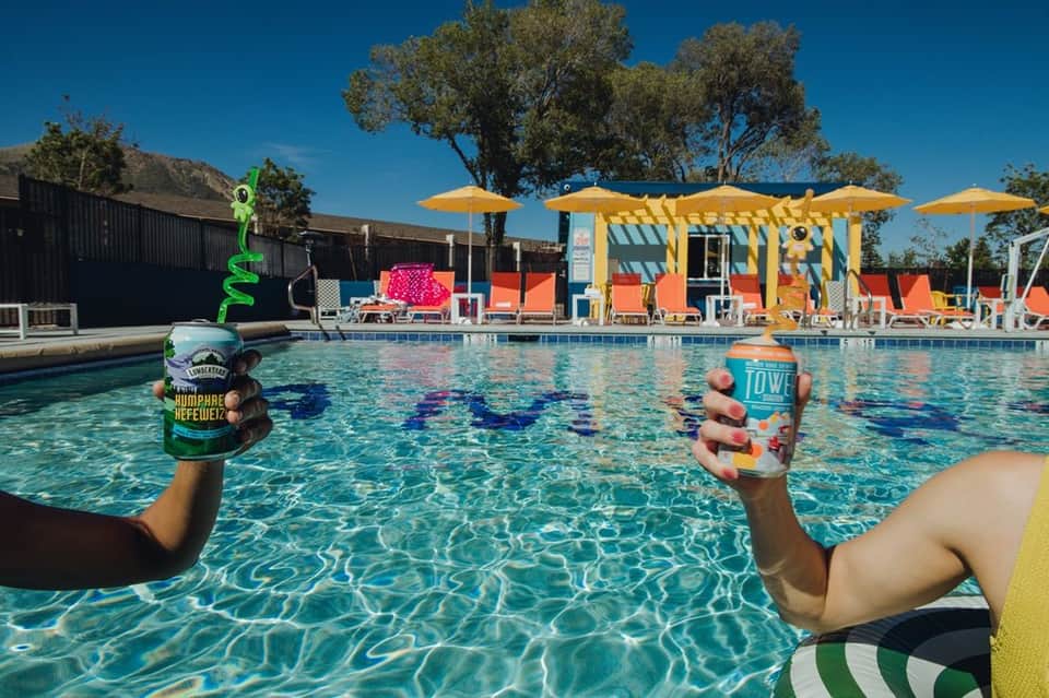 Two people in pool holding beverages with colorful pool toys and yellow pergolas with orange loungers in background
