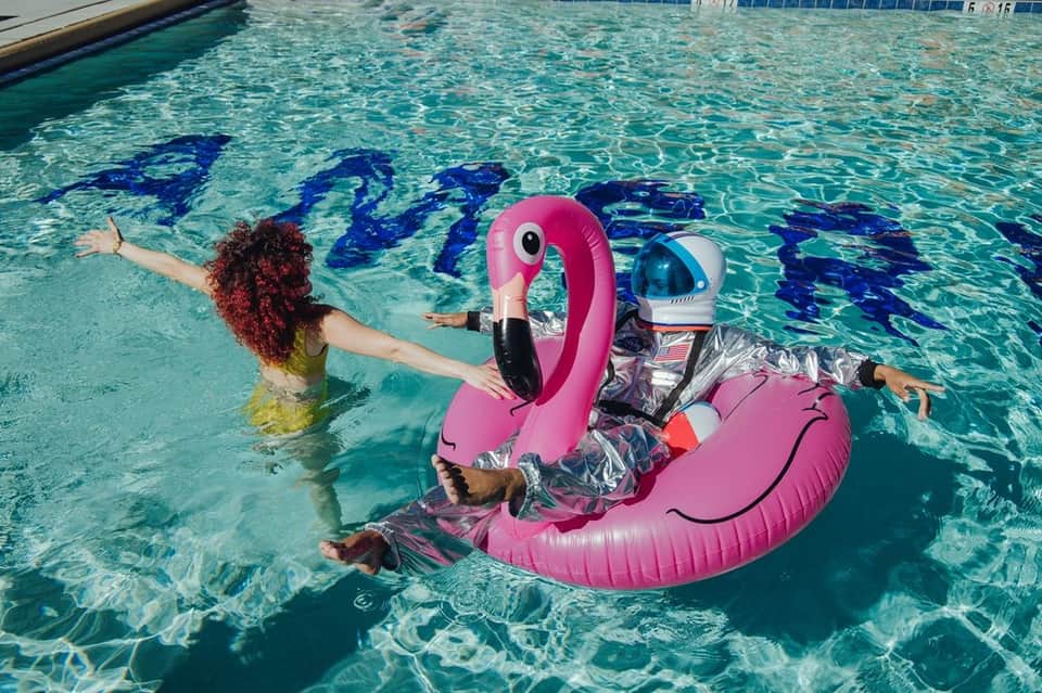 Woman with curly red hair on large pink flamingo pool float in clear turquoise water with blue lane dividers visible