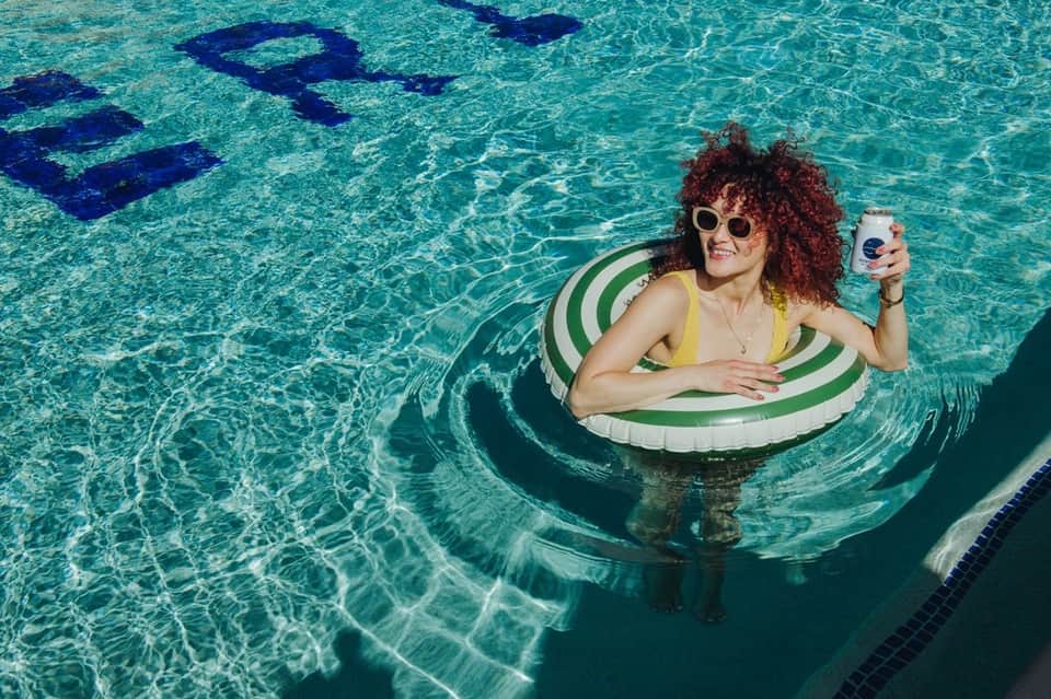 Woman in yellow swimsuit floating on green and white striped ring holding beverage in crystal clear pool water