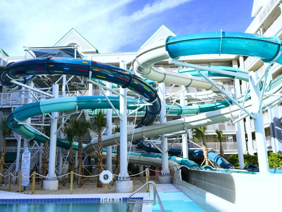 Complex water slide structure with turquoise and blue slides against white resort building with multiple balconies