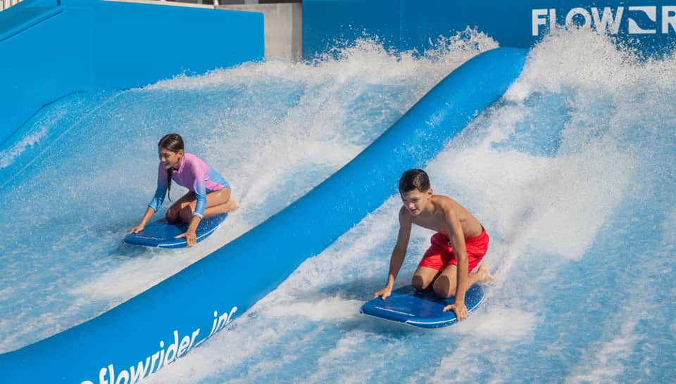 Two children surfing on FlowRider wave simulator on blue artificial wave with white water spray