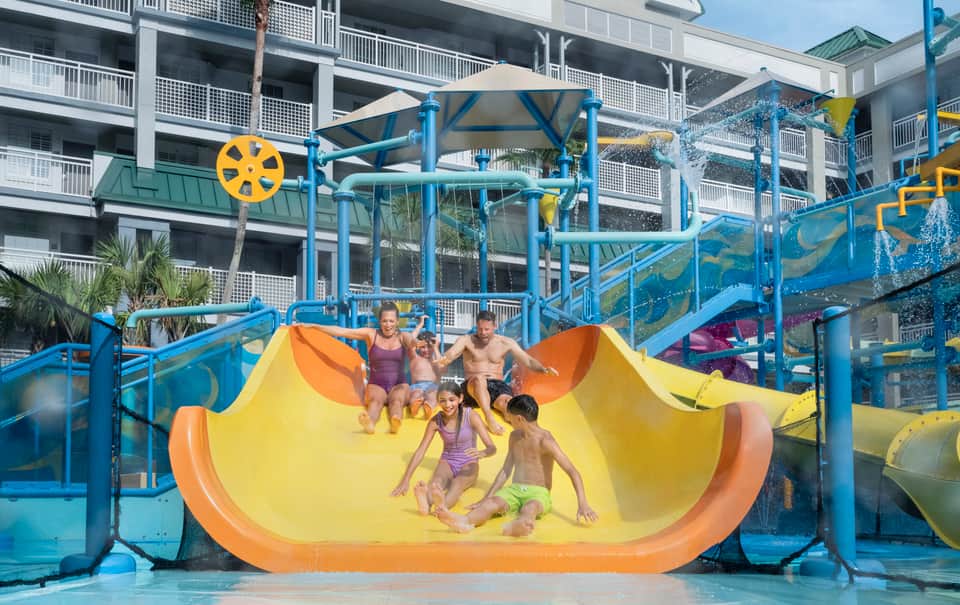Family of five riding large yellow waterslide together at resort with blue support structure and resort building