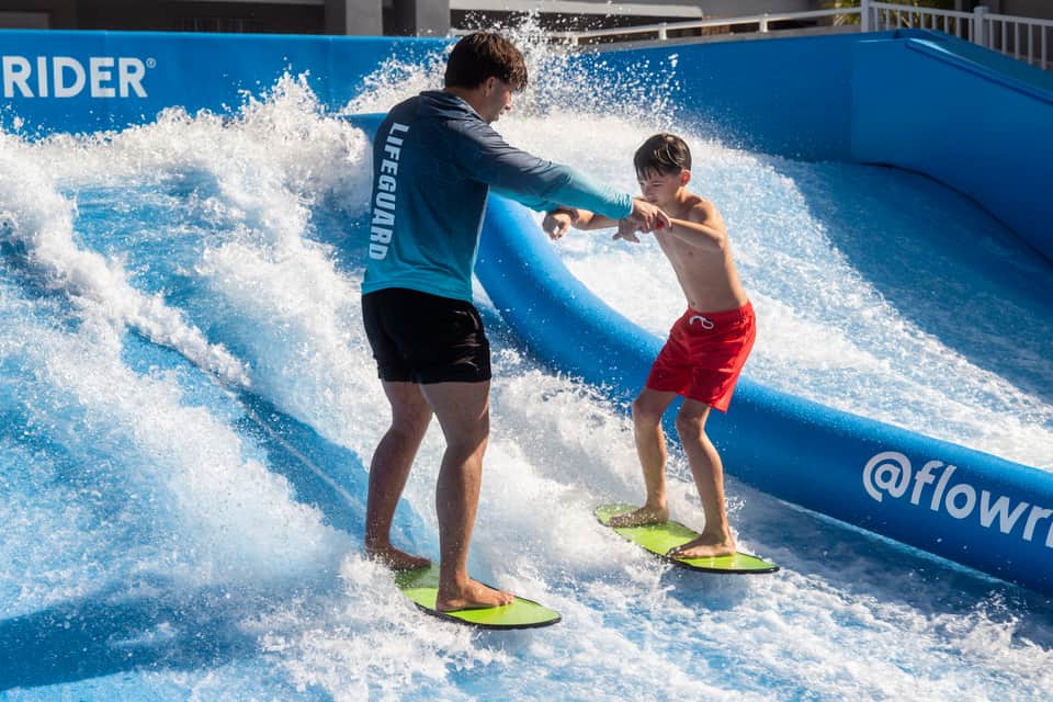 Instructor teaching young boy to surf on blue FlowRider wave with white water spray