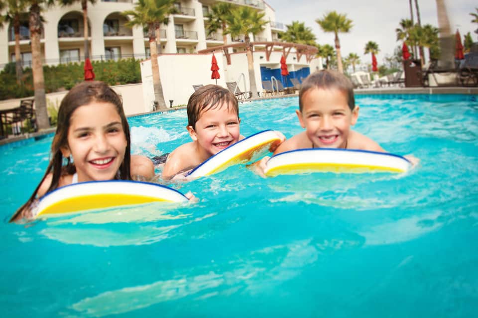 Three children enjoying swimming in a bright blue resort pool with kickboards