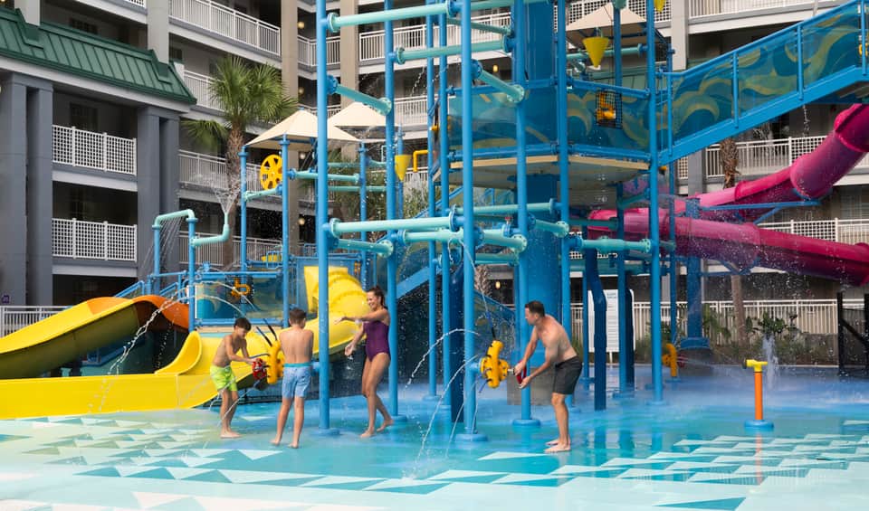 Group of children playing on blue water play structure with yellow and pink slides at resort splash pad