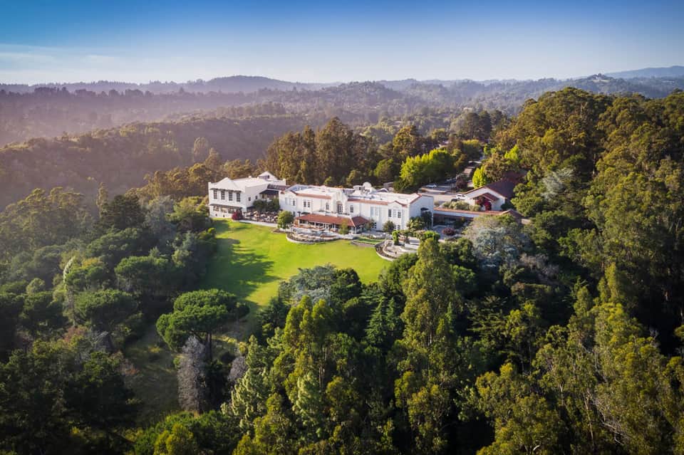 Aerial view of luxurious white mansion nestled in forested hillside with manicured lawns