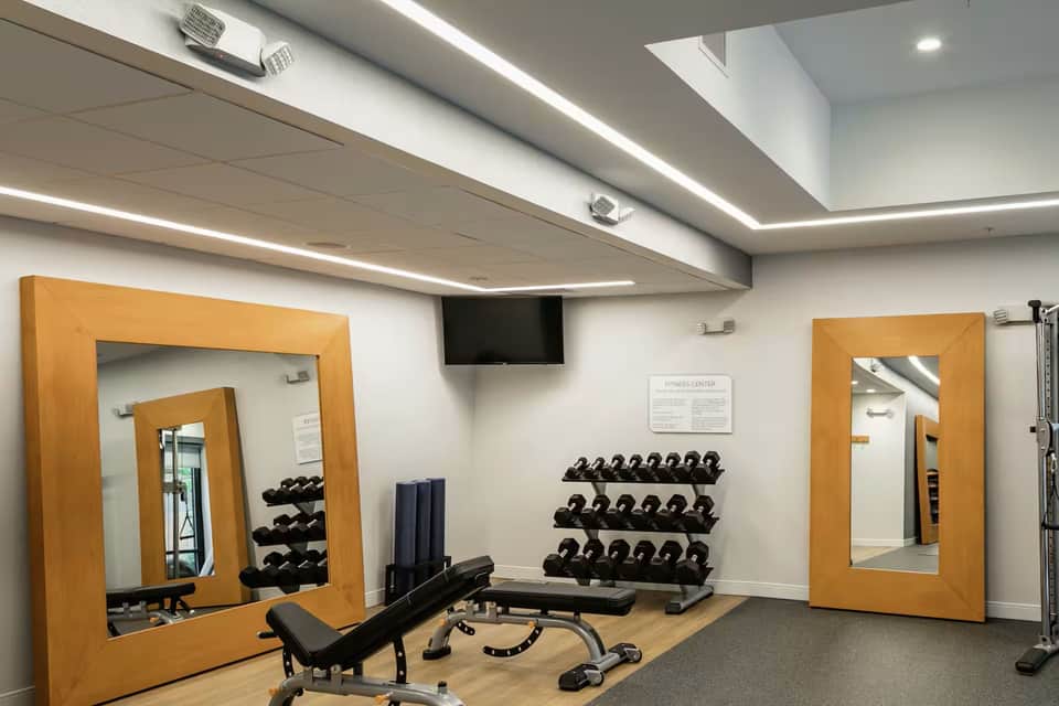 Hotel fitness center with mirrored walls, dumbbell racks, exercise equipment, and recessed ceiling lighting