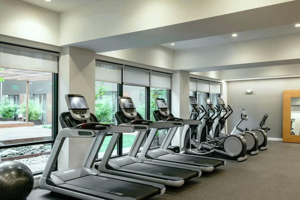 Modern fitness center with rows of treadmills and exercise equipment facing large windows overlooking gardens