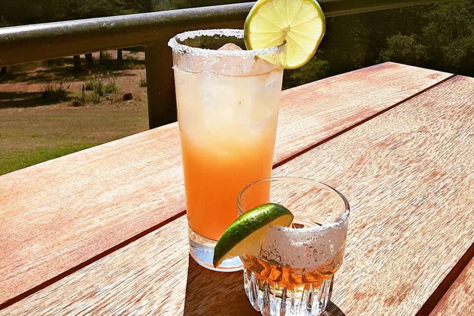 Two colorful cocktails on wooden deck with lemon and lime garnishes overlooking landscape