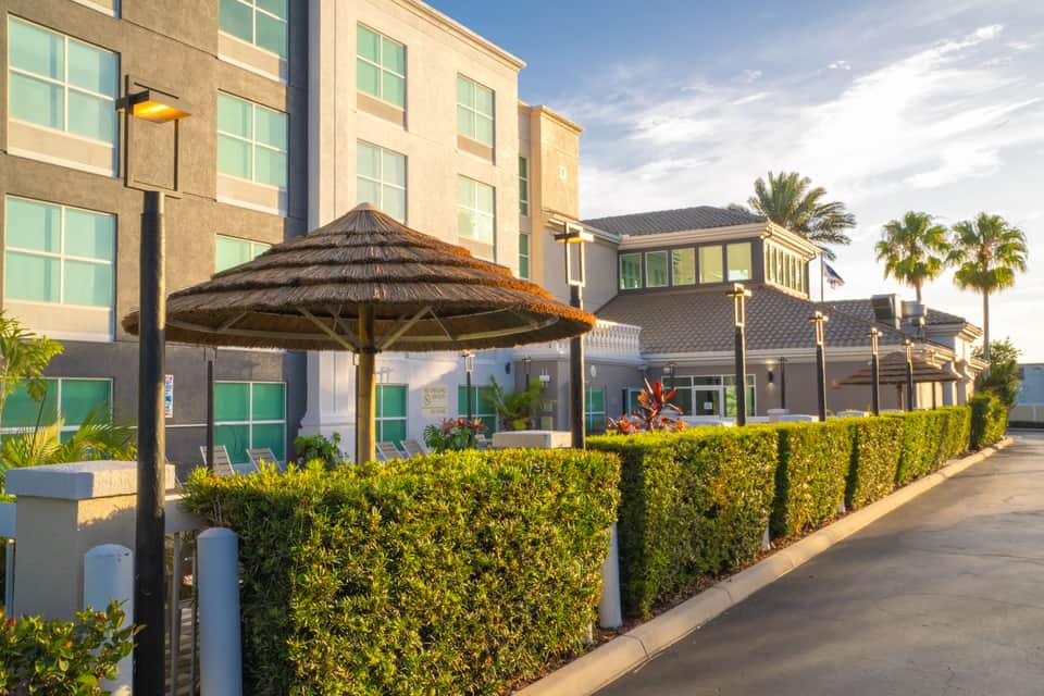 Modern hotel exterior with multi-story building, tropical landscaping, and thatched umbrella seating area
