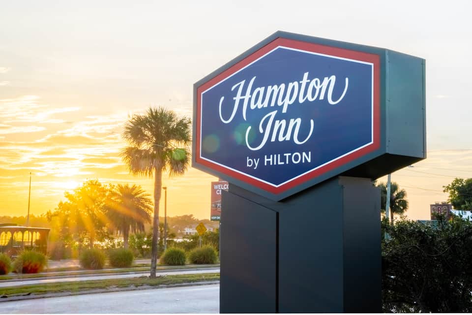Hampton Inn by Hilton sign at sunset with palm trees and entrance road