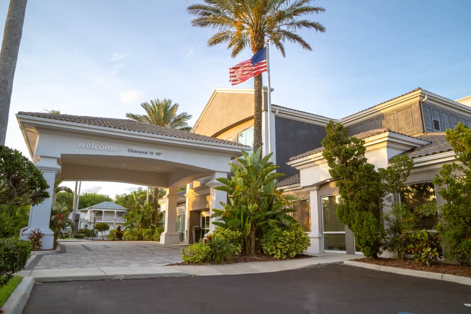 Hotel entrance with welcome canopy, American flag, palm trees, and manicured landscaping