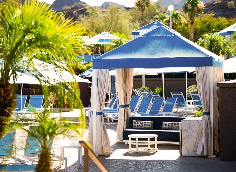 Resort pool area with blue cabanas, white lounge chairs, palm trees, and mountain backdrop