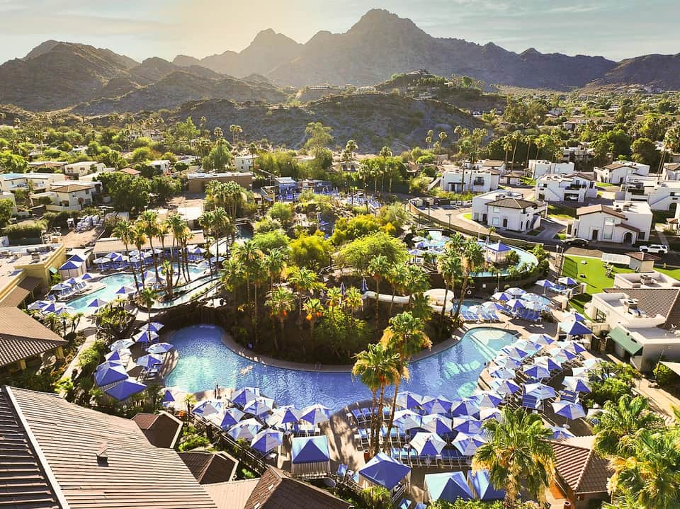 Aerial view of sprawling resort with multiple pools, blue umbrellas, palm trees, and desert mountain landscape