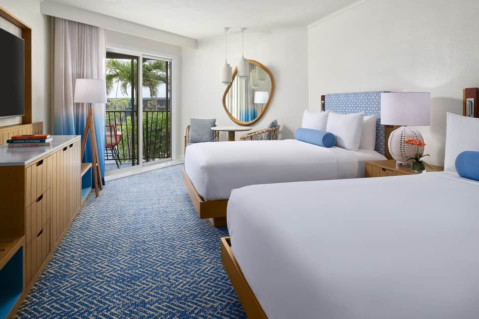 Modern hotel bedroom with two beds, ocean view balcony, blue accents, and wooden furniture