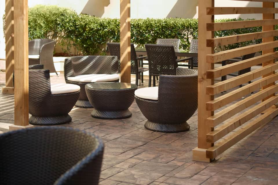 Patio seating area with woven chairs, white cushions, wooden pergola, and hedged privacy screening