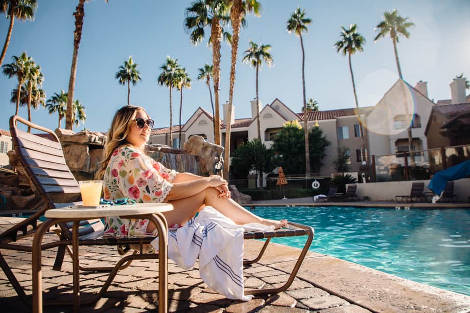 Woman in floral shirt relaxing poolside on lounge chair with palm trees and resort buildings in background