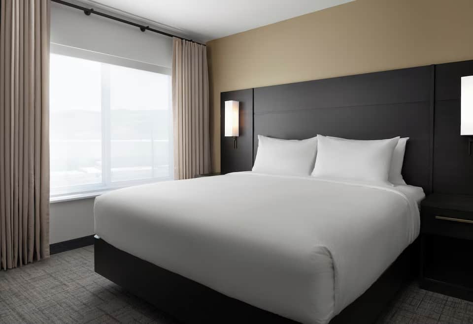 Modern hotel bedroom with king bed, black headboard, beige walls, and floor-to-ceiling windows with city views