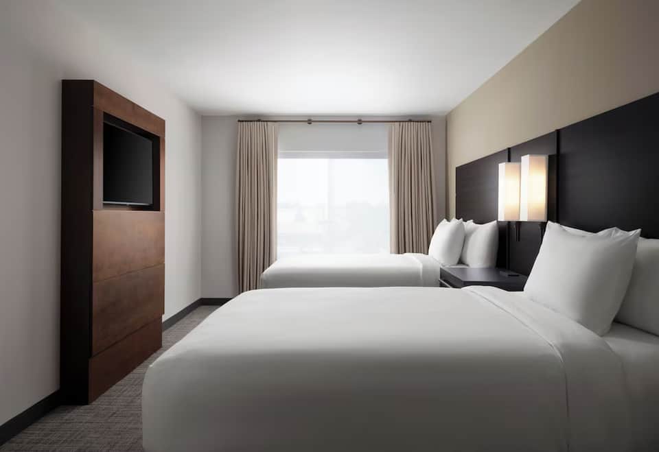 Hotel room with two queen beds, dark headboards, wall-mounted TV, window with curtains, and contemporary furnishings