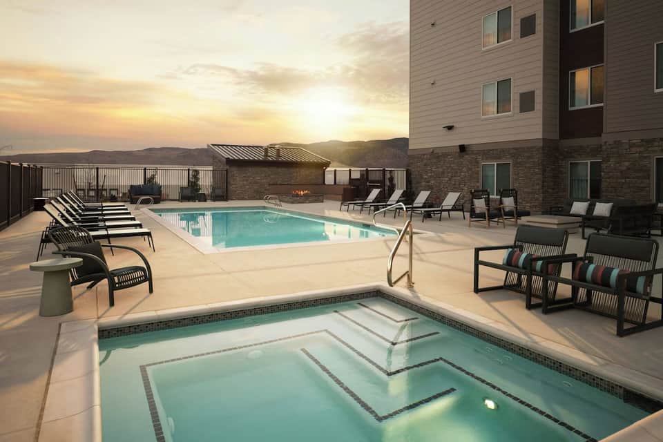 Outdoor pool deck at sunset with lounge chairs, hot tub, fire pit, and modern residential building backdrop