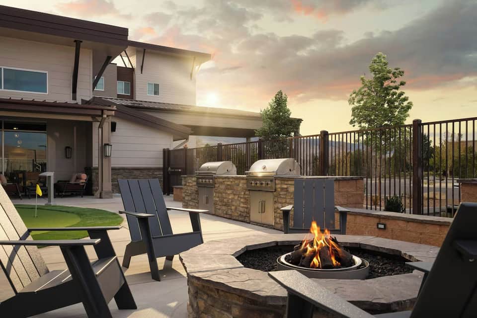 Backyard patio with fire pit, lounge chairs, stone grill counter, and modern house at golden hour