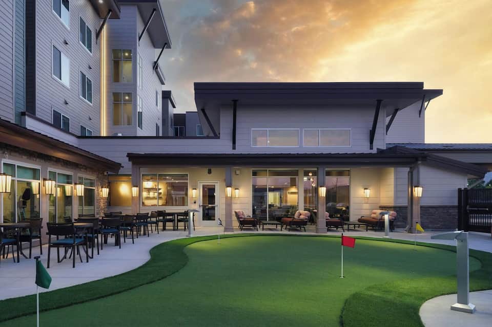 Upscale residential patio with dining area, lounge space, mini golf course, and illuminated building at dusk