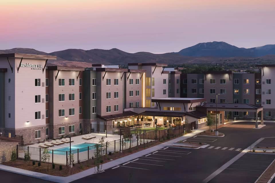 Residential complex at twilight with illuminated lobby, pool area, and mountain backdrop against pink sky