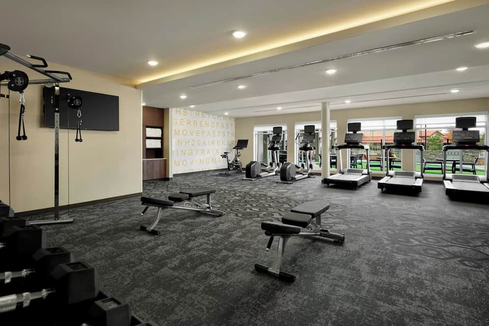 Modern fitness center with cardio equipment, strength training benches, TV screens, and large windows with views