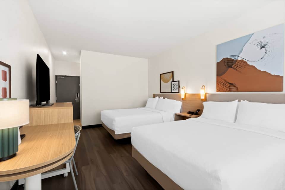 Hotel room with two queen beds, wooden furniture, abstract wall art, and TV
